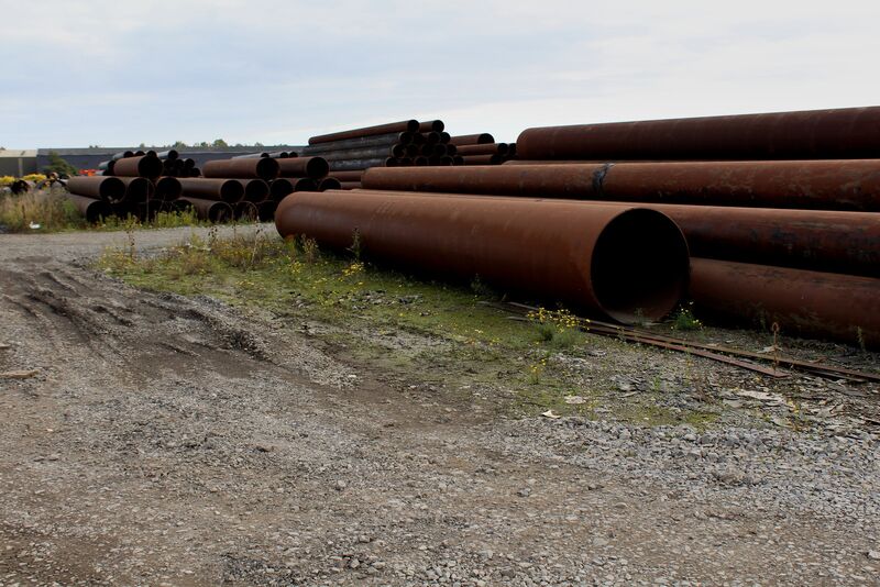Large rusty steel pipes lying on gravel, with stacks of smaller pipes behind them